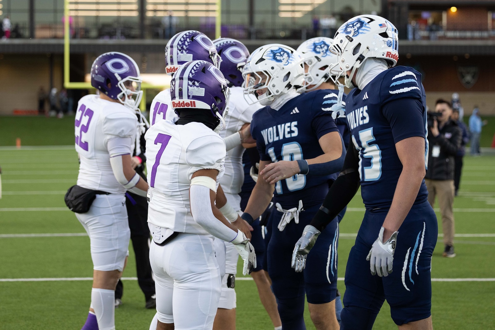 CHS-WPHS-FB-6 | Press Pass Sports Football players from a white and purple team shake hands with blue-clad Wolves on a green field before kickoff, helmets on.
