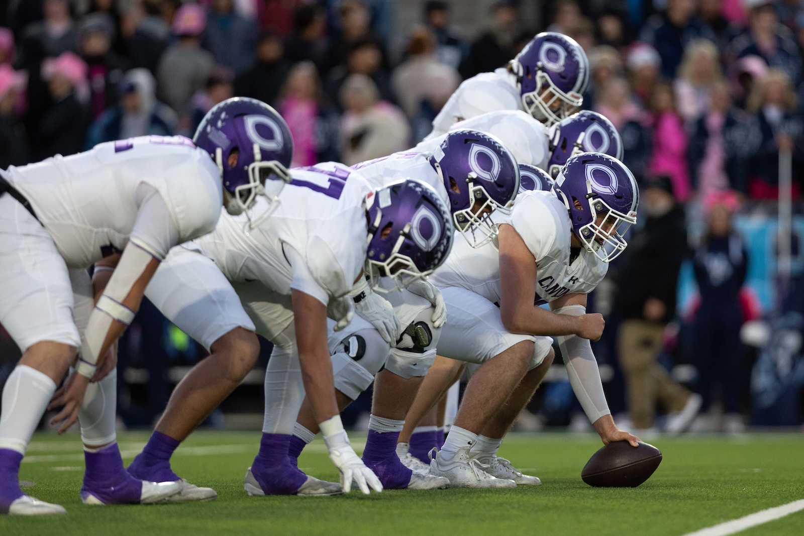 CHS-WPHS-FB-12 | Press Pass Sports Football team lined up at the snap, center about to snap the ball, white uniforms with purple helmets, on a green field with a crowd in the background.