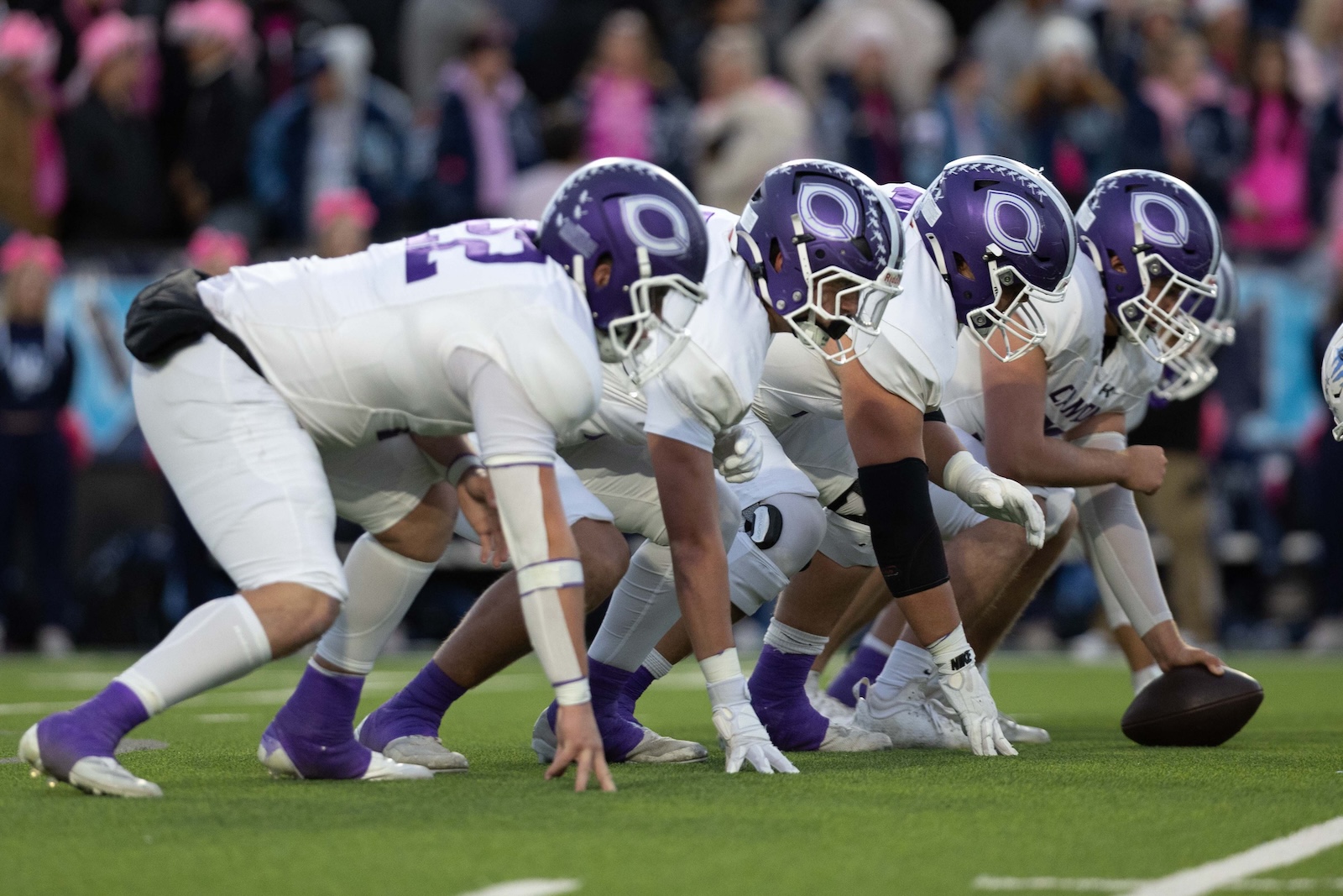 CHS-WPHS-FB-13 | Press Pass Sports American football players in white uniforms crouch at the line of scrimmage, purple helmets ready for snap with a crowd in the background, on a green field.