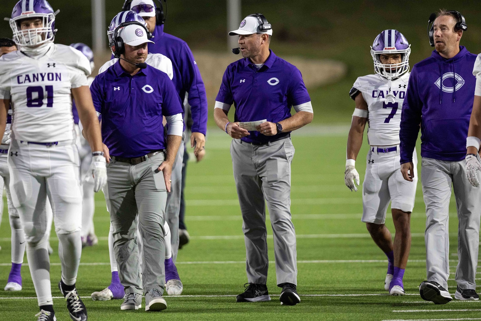 CHS-WPHS-FB-17 | Press Pass Sports Coaches and players in purple and white Canyon football uniforms on a green field, some wearing headsets and a coach holding a play sheet.