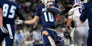 Football player wearing navy jersey #10 runs with the ball as teammates and defenders battle nearby on the field
