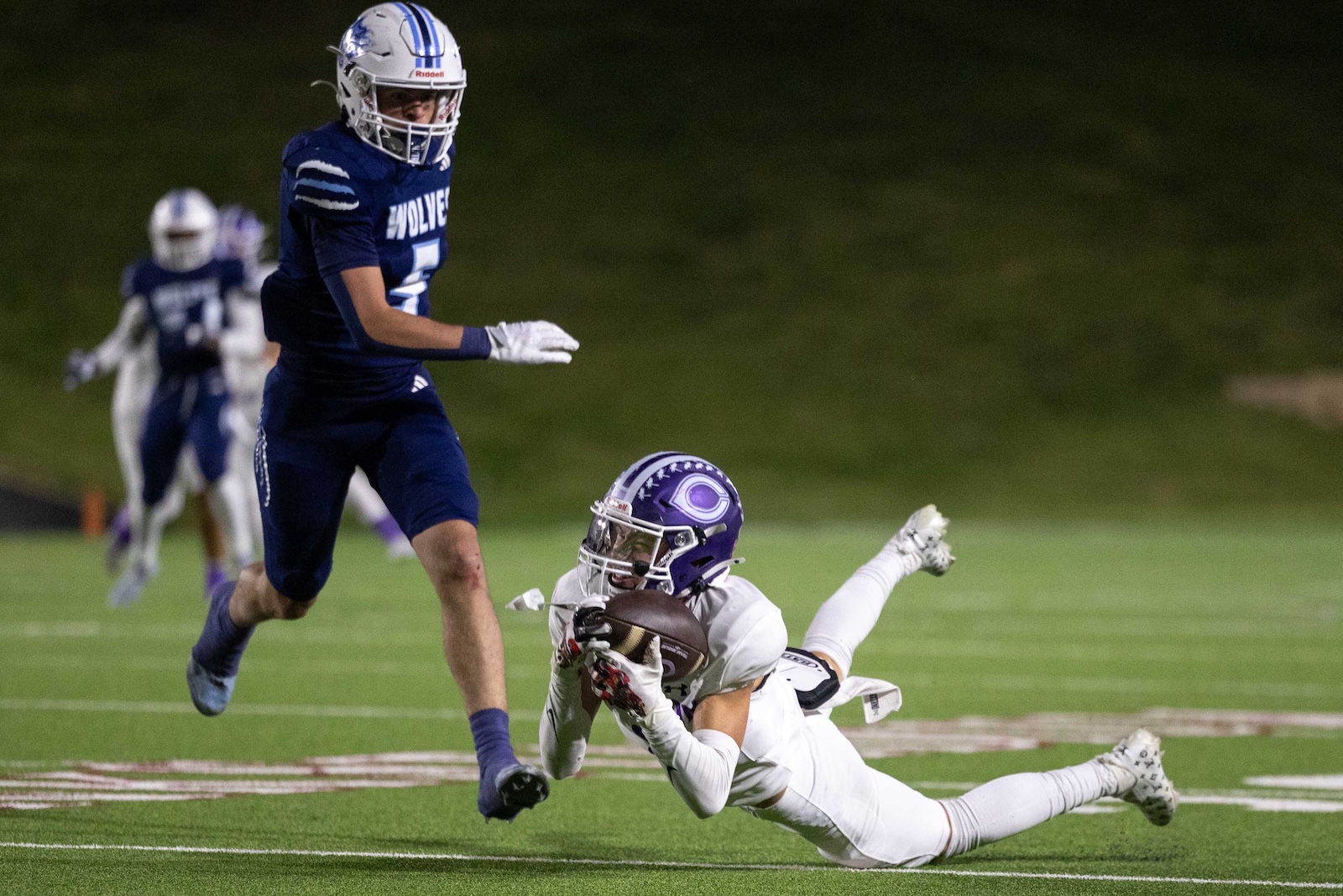 CHS-WPHS-FB-26 | Press Pass Sports Night football game: a blue-uniformed player with the ball runs forward while a white-uniformed defender with a purple helmet dives to tackle him on the turf.
