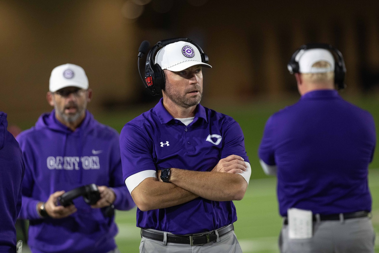 CHS-WPHS-FB-27 | Press Pass Sports Three coaches in purple shirts and white caps stand on a field at night, wearing headsets and talking.”
