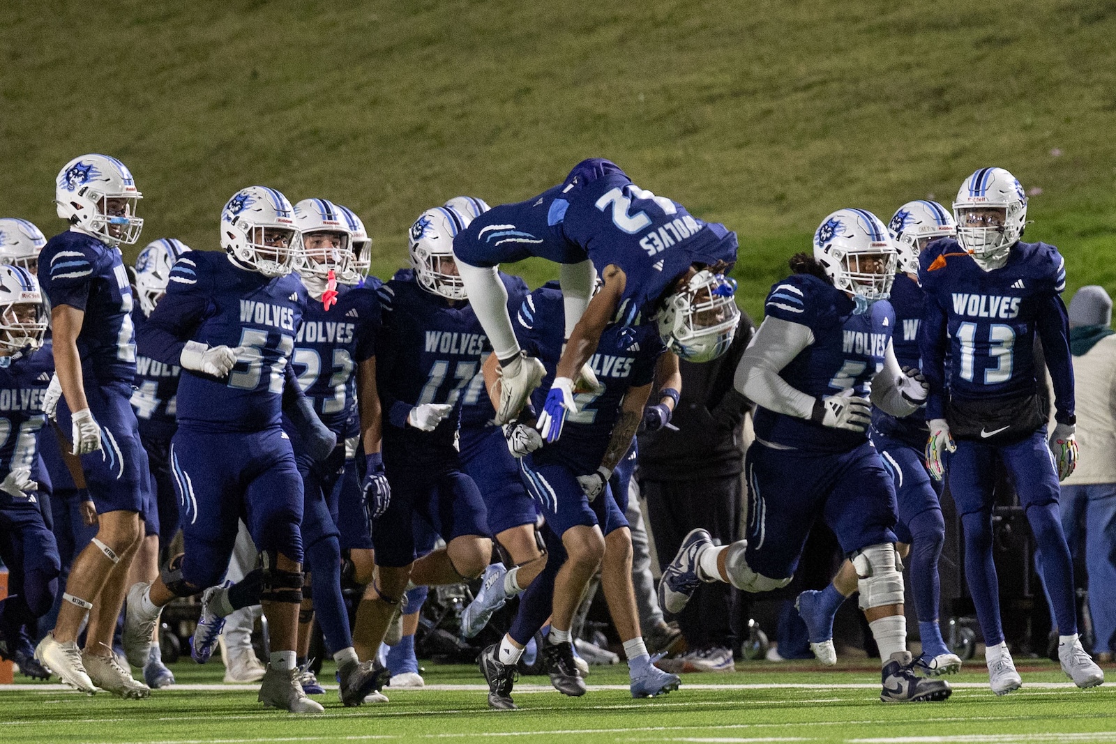 CHS-WPHS-FB-28 | Press Pass Sports Wolves football team in blue uniforms huddle on the field as a player leaps midair over teammates during a celebration moment.