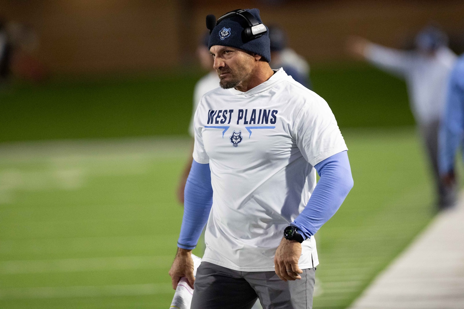CHS-WPHS-FB-32 | Press Pass Sports Coach on a sports field wearing a white West Plains shirt and blue sleeves, headset, and beanie, looking focused as he walks along the sideline.
