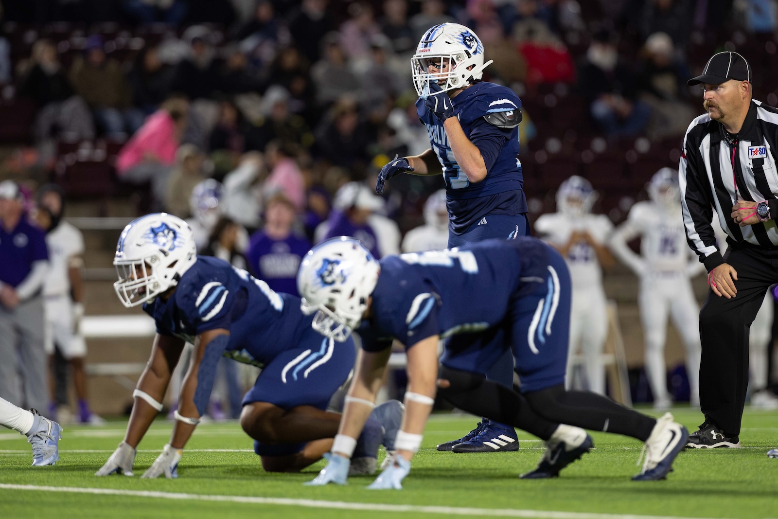CHS-WPHS-FB-33 | Press Pass Sports Football players in blue uniforms line up on the 40-yard line as a quarterback jumps to signal a play, with a referee on the right and blurred crowd in the background.