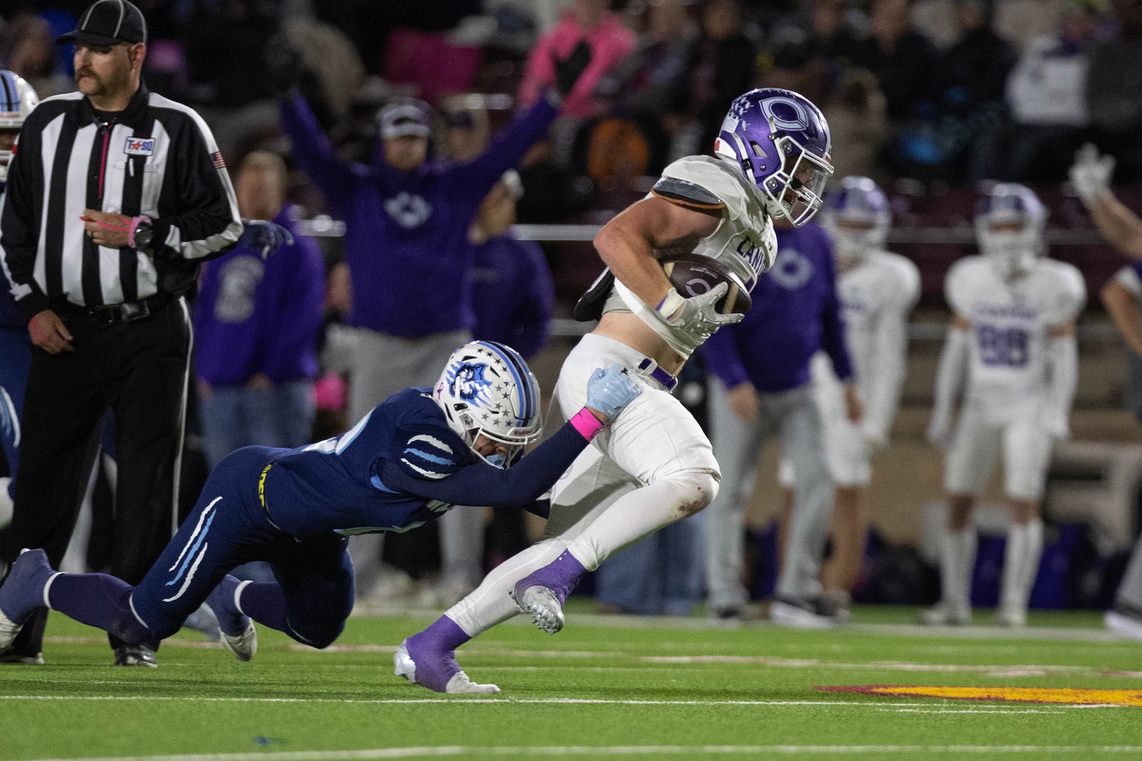 CHS-WPHS-FB-35 | Press Pass Sports Football game action: a white-purple team player runs with the ball as a blue-team defender dives in for a tackle with a referee nearby.