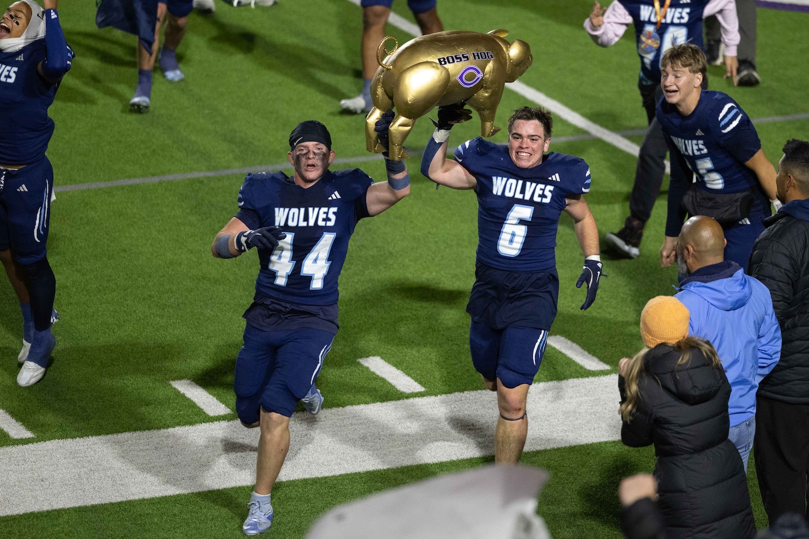 CHS-WPHS-FB-47 | Press Pass Sports Two Wolves football players lift a gold hog-shaped trophy on the field as teammates celebrate nearby in blue uniforms.