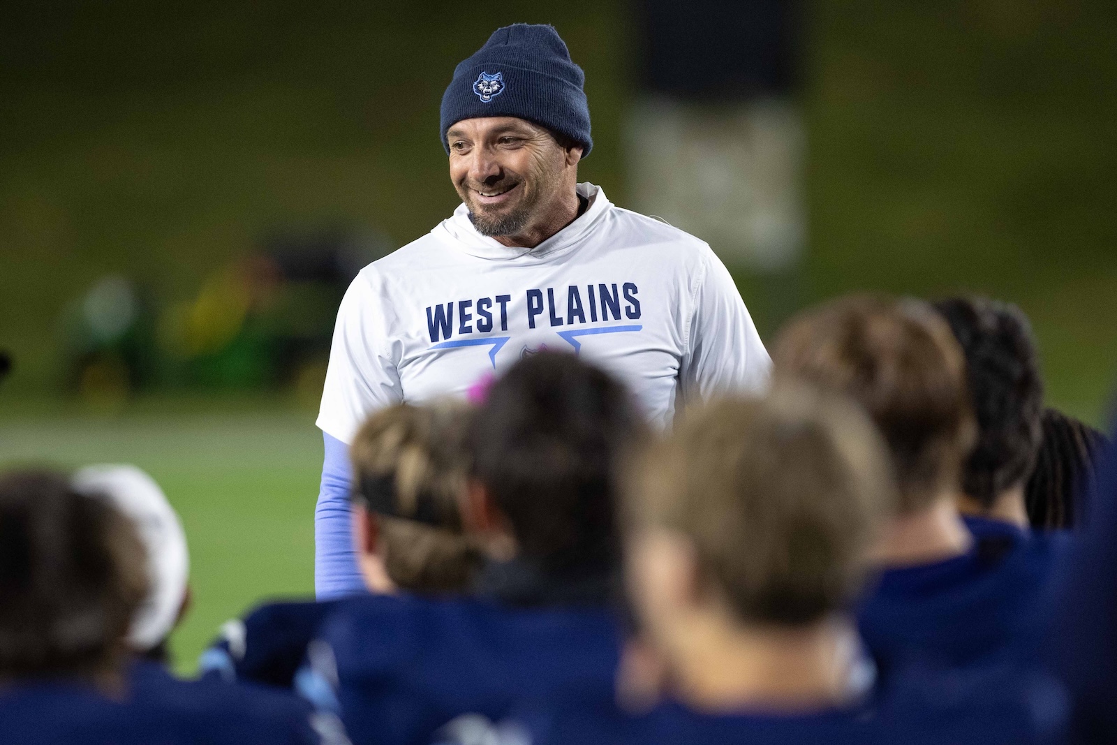 CHS-WPHS-FB-51 | Press Pass Sports Coach smiling in a white West Plains shirt addressing players during a huddle on a field at night, wearing a navy beanie.