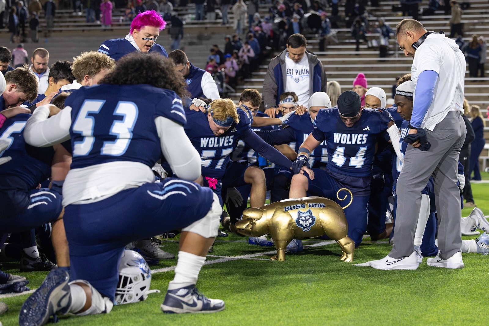 CHS-WPHS-FB-52 | Press Pass Sports Blue football team kneels in a circle around a gold pig statue labeled'Boss Hog' on the field, hands joined in a moment of unity before a game.