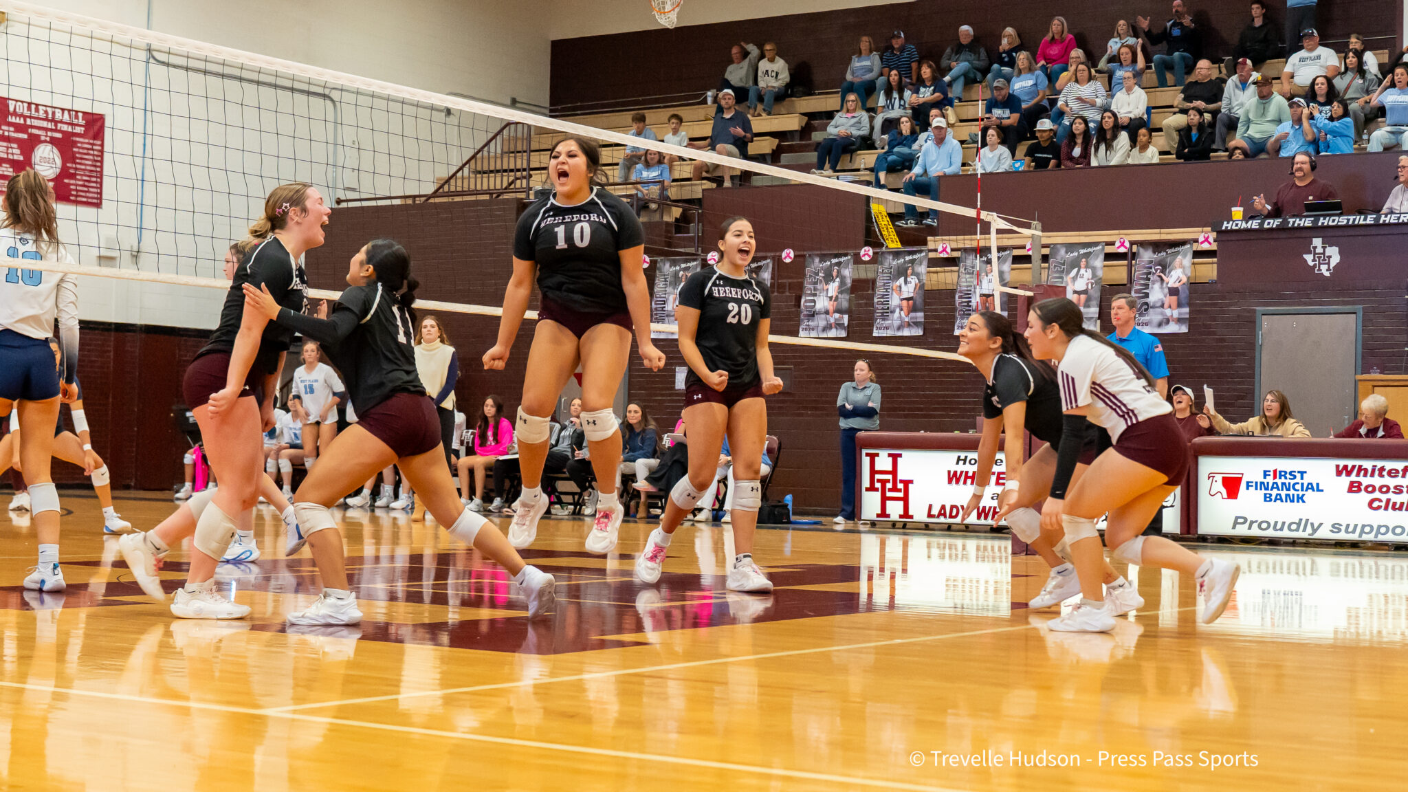 DSC08271 | Press Pass Sports Celebrating volleyball players on the court near the net after a point, with cheering teammates and spectators in the bleachers behind them.