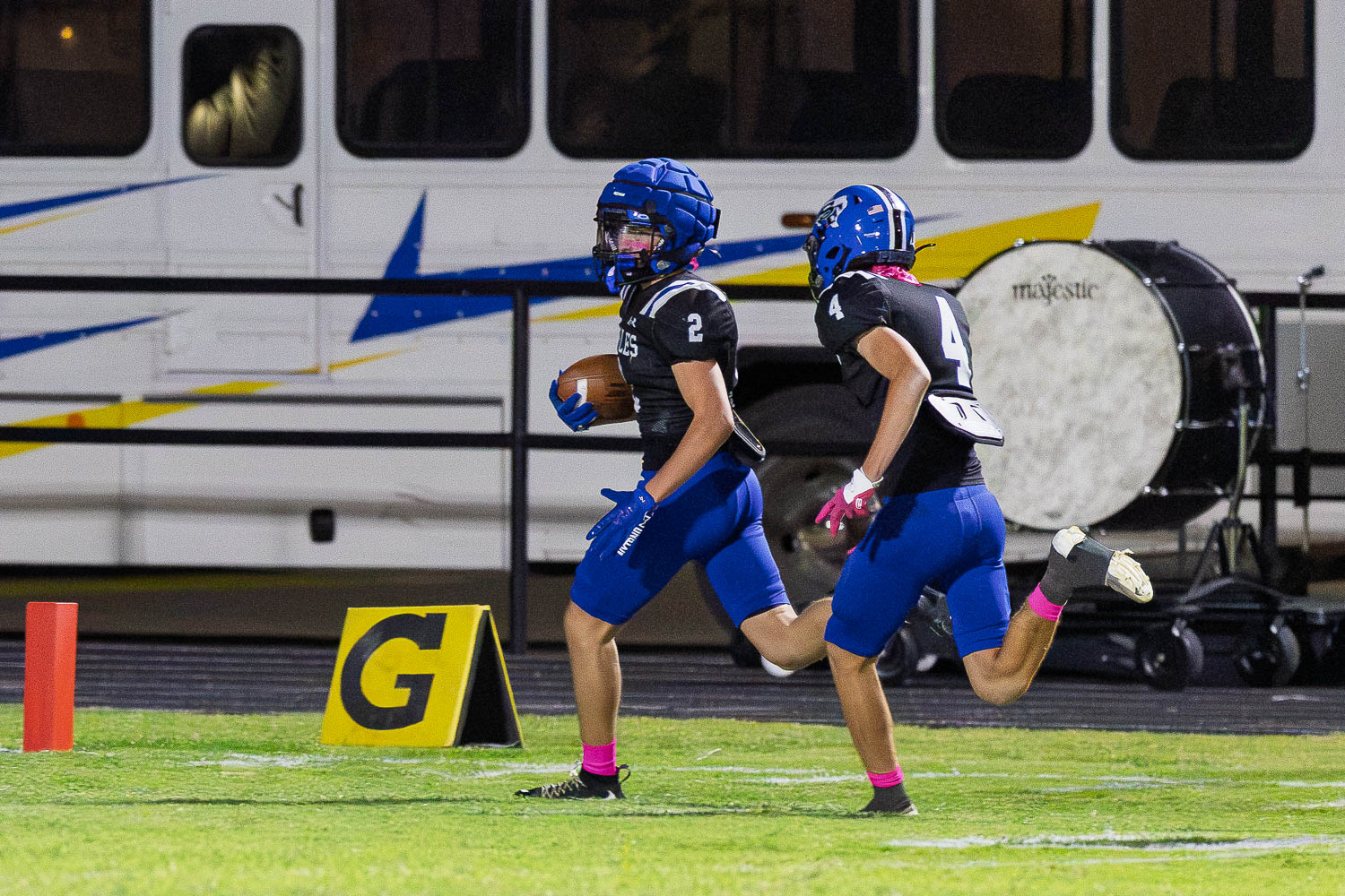 Two football players in blue and black uniforms sprint toward the end zone, one carrying a football, as a large drum and a bus stand in the background.