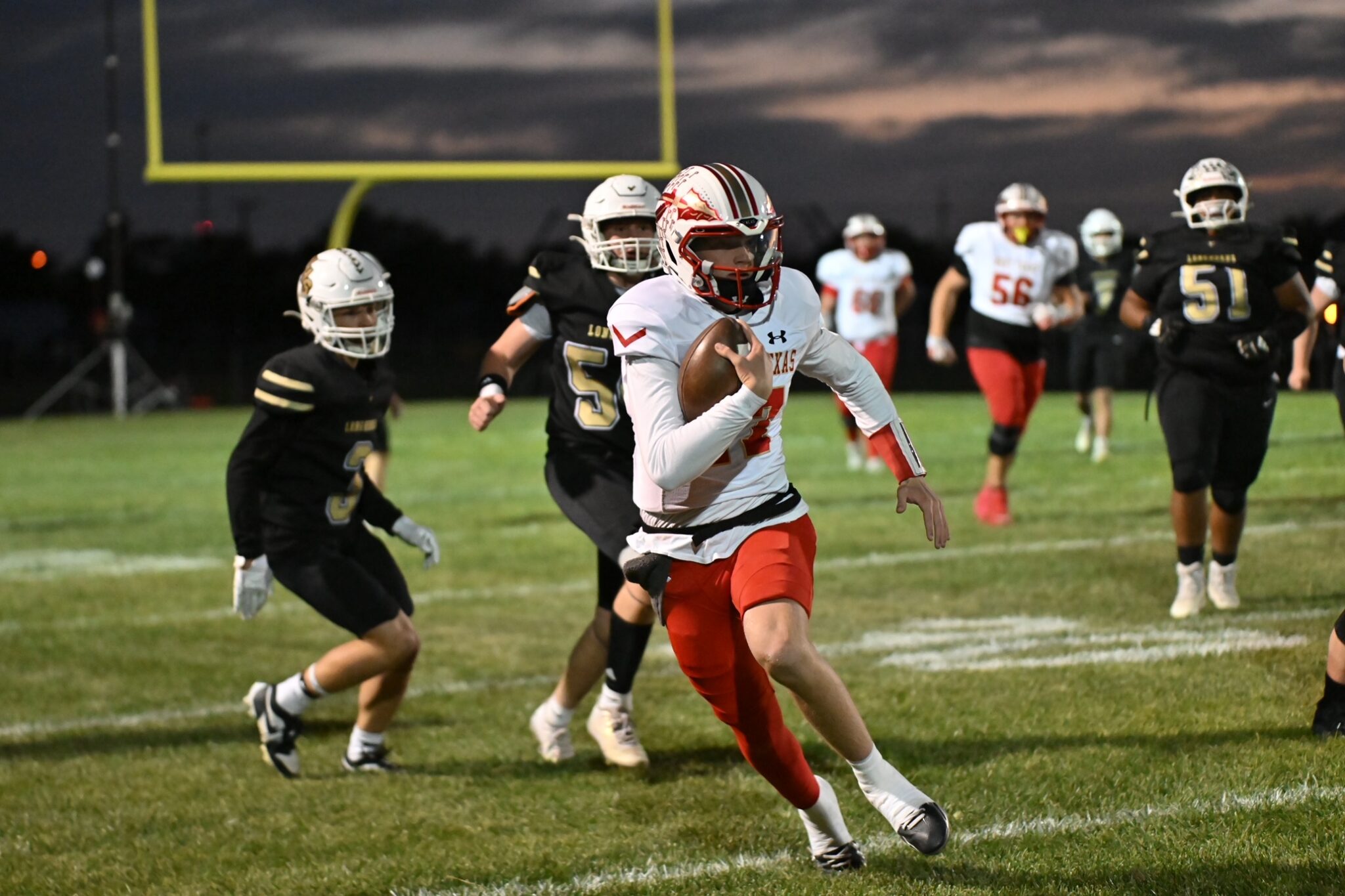 image7 3 | Press Pass Sports Football player in white and red jersey sprints with the ball as defenders in black chase him on a grassy field at dusk; a yellow goalpost is in the background.