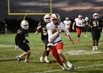 image7 3 | Press Pass Sports Football player in white and red jersey sprints with the ball as defenders in black chase him on a grassy field at dusk; a yellow goalpost is in the background.