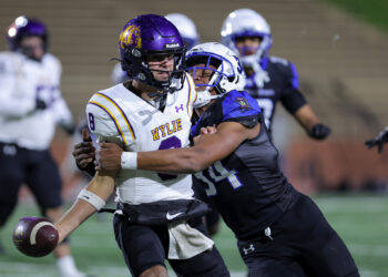 Two football players collide on the field during a tackle: a white-uniformed player with a purple 'WYLIE' helmet holds the ball as a dark-shirted defender grabs him.