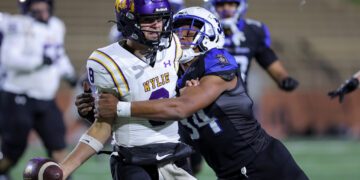 Two football players collide on the field during a tackle: a white-uniformed player with a purple 'WYLIE' helmet holds the ball as a dark-shirted defender grabs him.