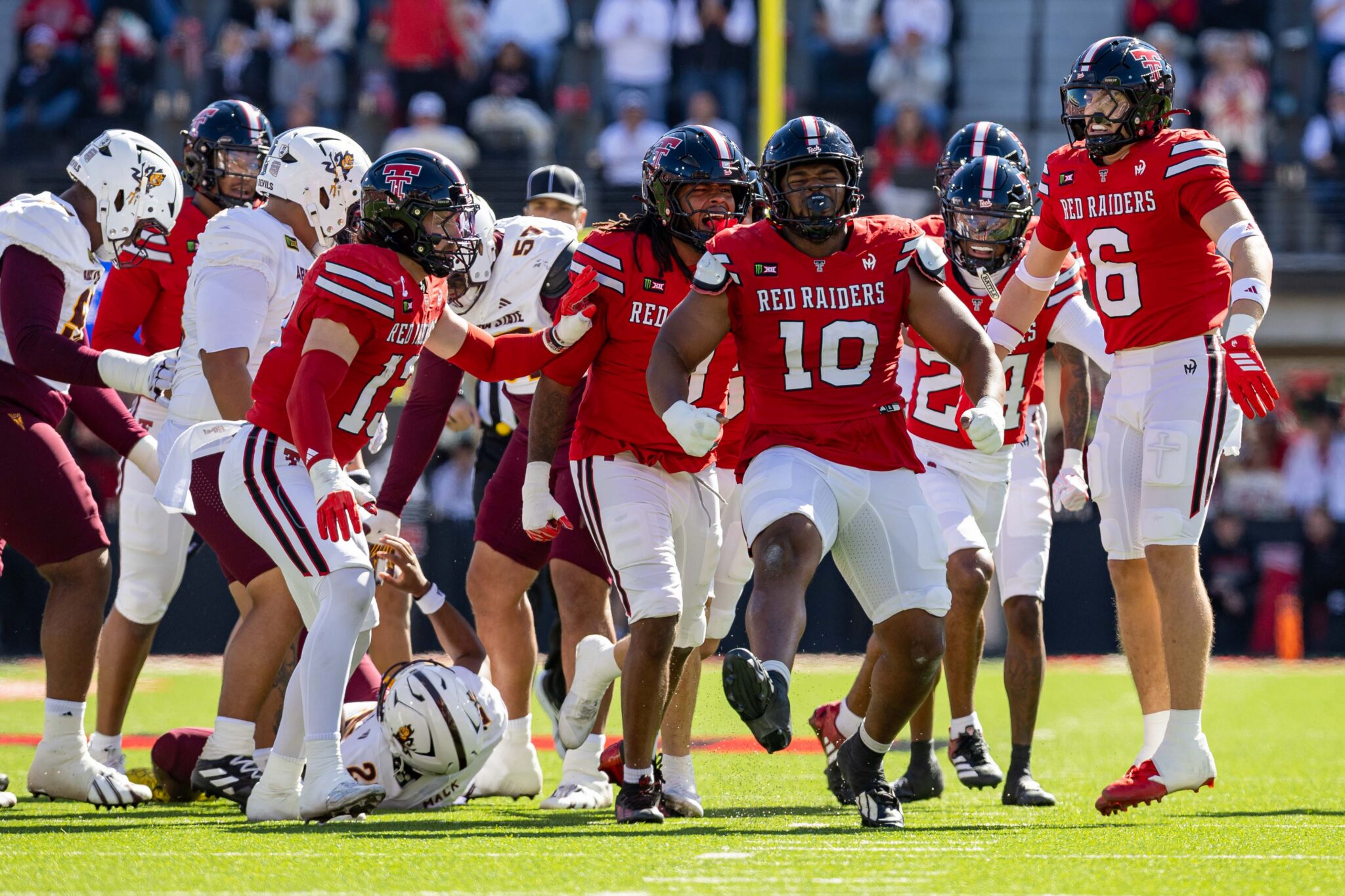 Red Raiders players in red jerseys celebrate on a sunny football field, with a fallen ball at their feet and teammates embracing.