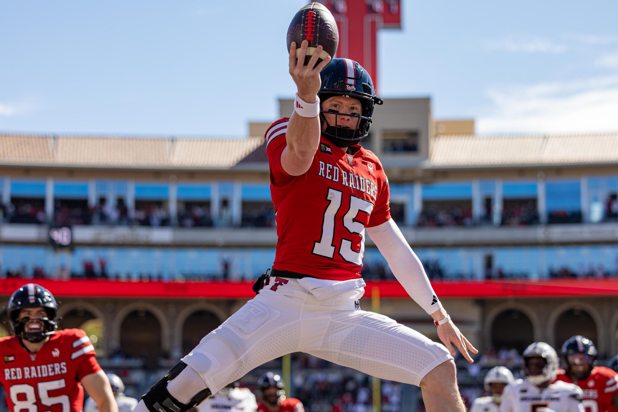 Football player in a red Red Raiders jersey leaps to catch a ball during a sunny game in a stadium.