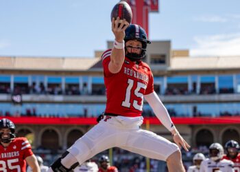 Football player in a red Red Raiders jersey leaps to catch a ball during a sunny game in a stadium.