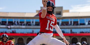 Football player in a red Red Raiders jersey leaps to catch a ball during a sunny game in a stadium.