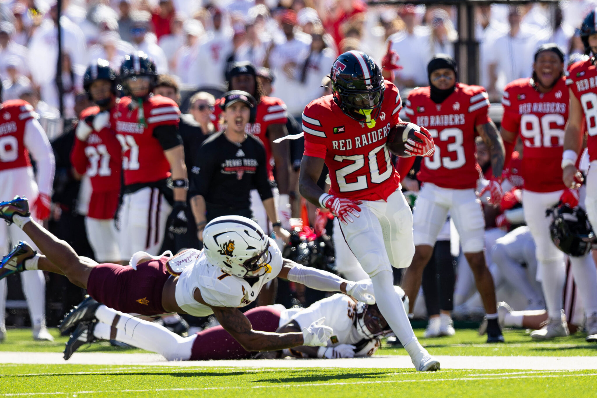 Red Raiders player in red jersey runs with the ball as defenders dive and fans cheer in the background on a sunny day.