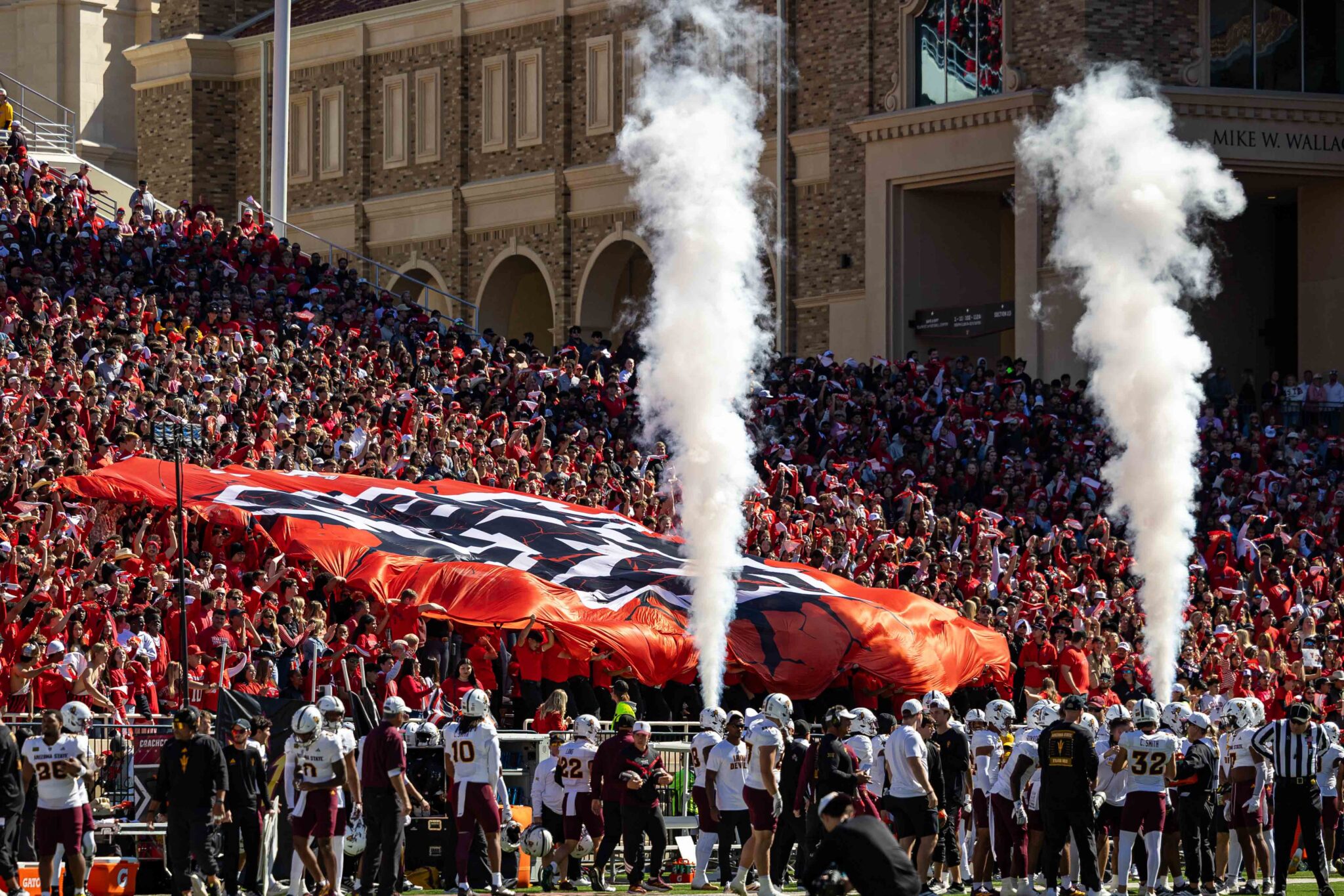 Crowd of football fans in red cheer in a stadium, with white smoke plumes rising from the field and a marching band at the bottom edge.