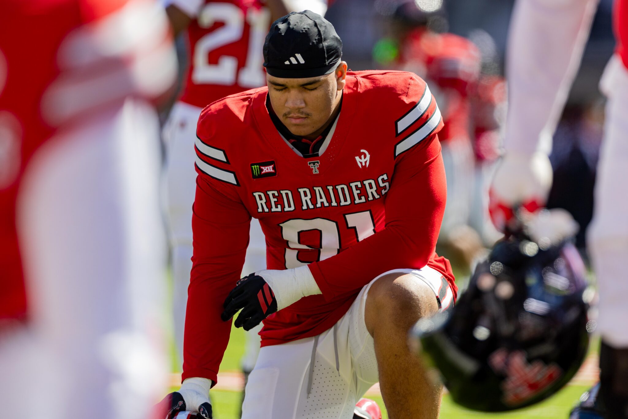 Close-up of a Red Raiders football player in a red jersey kneeling on the field, helmet in hand.
