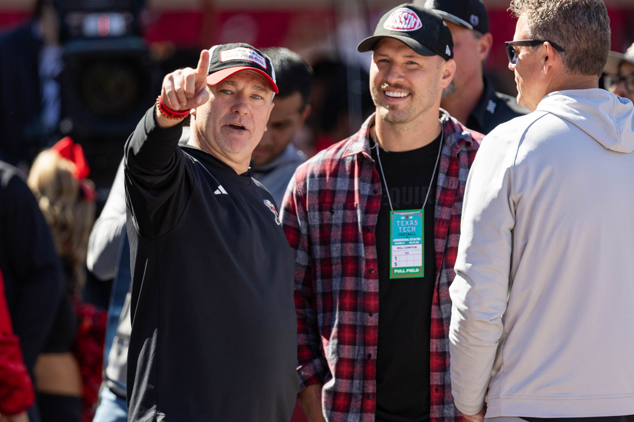 Man in a black shirt points toward the camera while talking with others at an outdoor event; a plaid-shirted man with a badge stands nearby.
