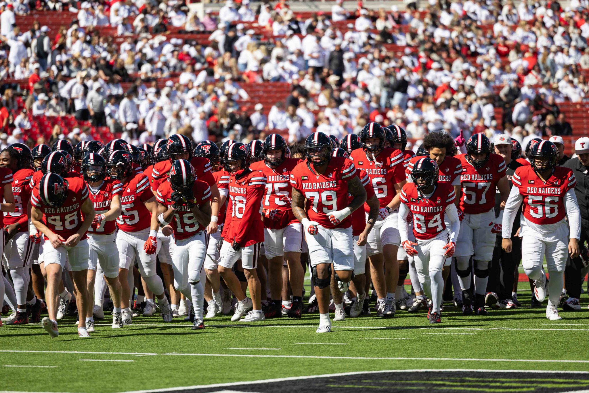 Red Raiders football team in red jerseys marching onto the field as a packed stadium applauds in the stands.