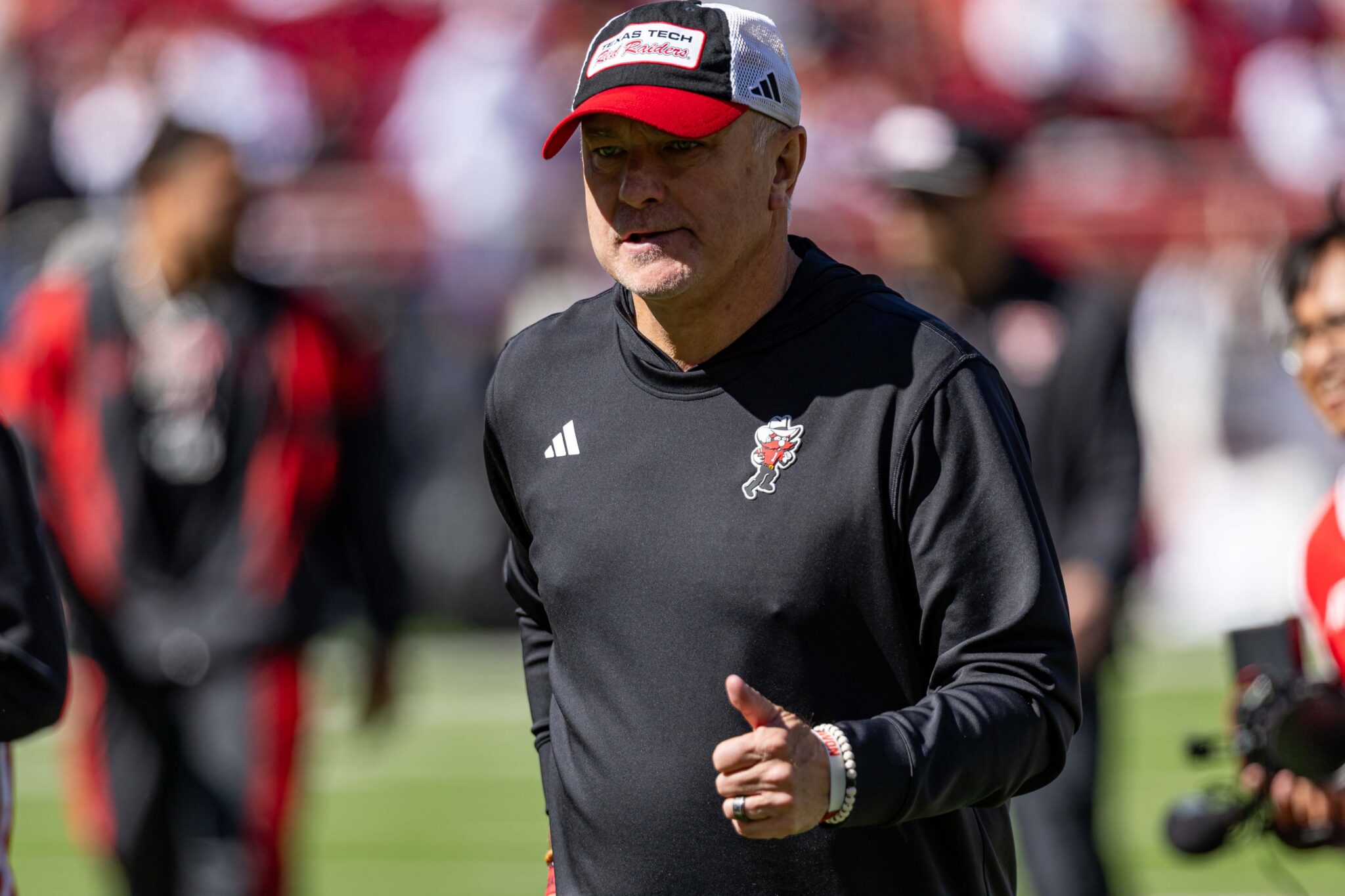 Man in a black athletic shirt runs on a grassy field, wearing a red-and-white cap with a team logo and an Adidas emblem on the chest.