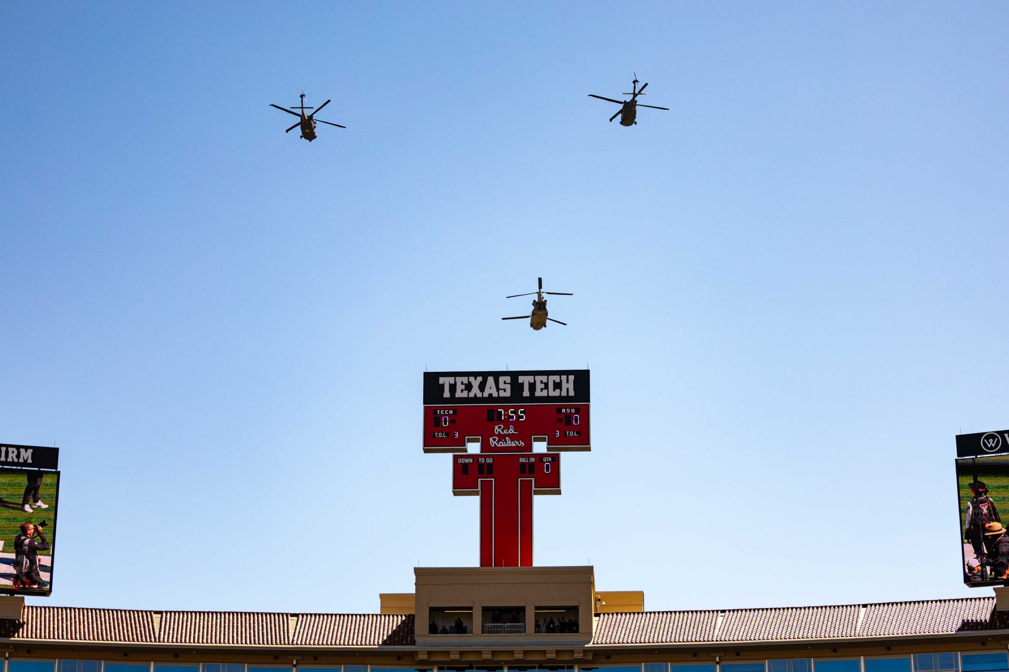 Three helicopters fly in formation above a Texas Tech stadium scoreboard against a clear blue sky, with large video boards on either side.