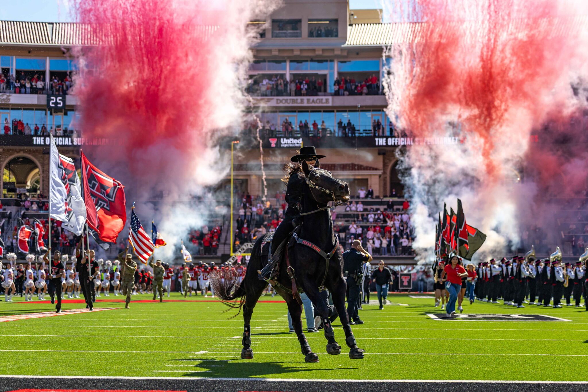 Mounted rider on a black horse leads a parade on a football field with red smoke filling the air and spectators in the stands.