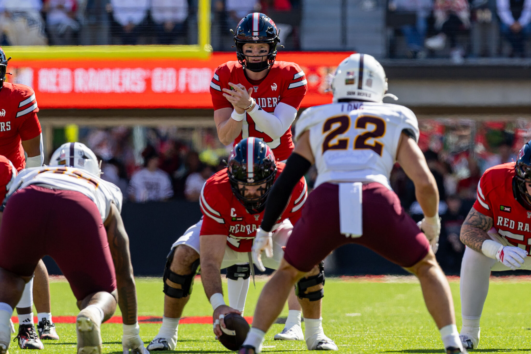 Red Raiders quarterback in a red jersey, ball in hand, preparing to pass while defenders converge during a college football game on a sunny field.
