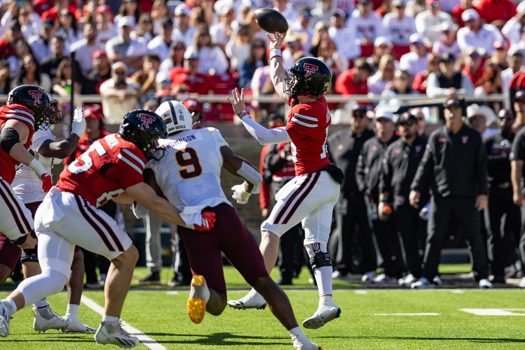 Football players in red and white tackle as a quarterback tosses the ball during a game with a crowded stadium backdrop.