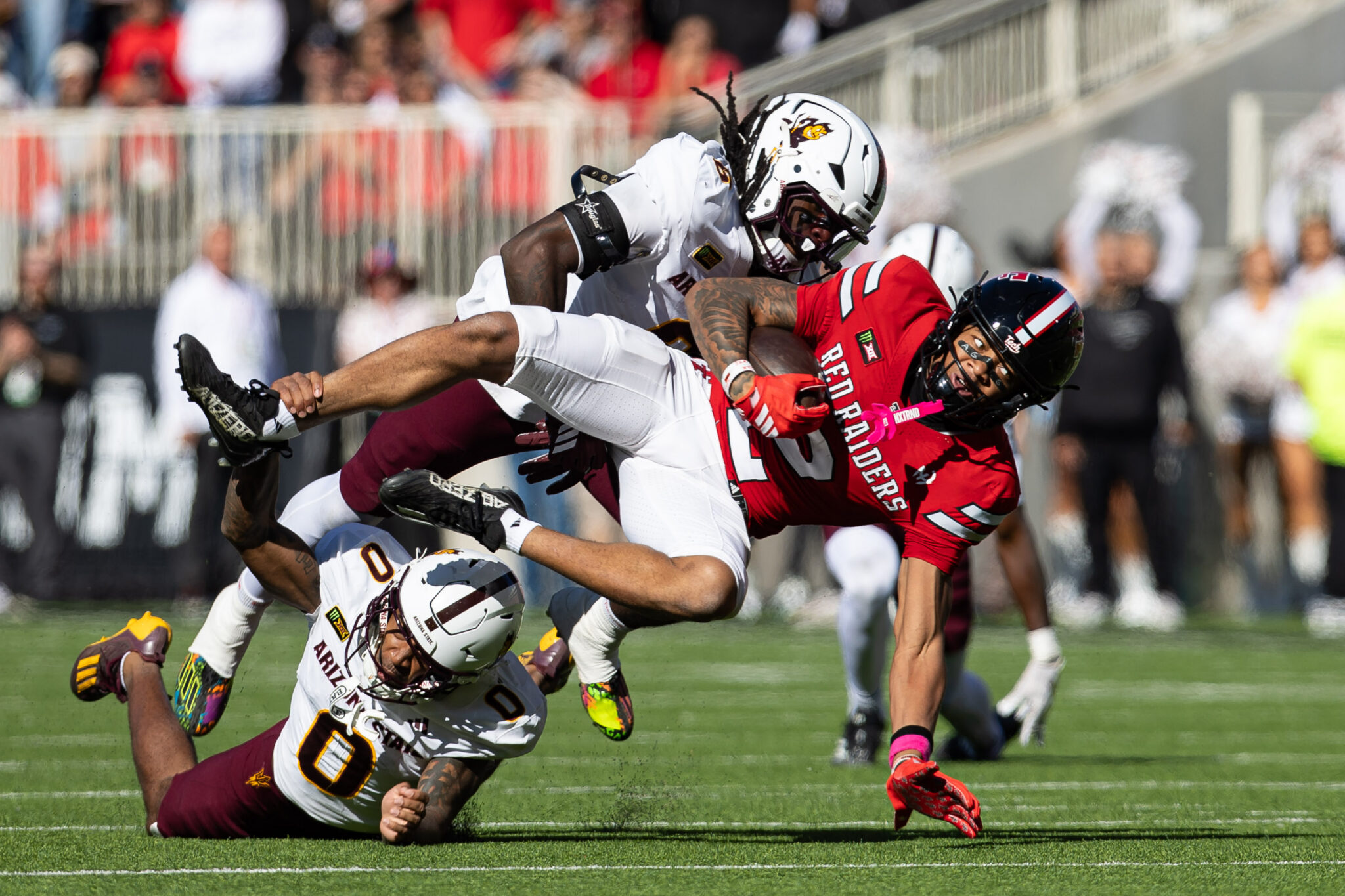 Two football players collide mid‑air during a tackle on a sunny field, wearing white and red uniforms with a blurred crowd in the background.