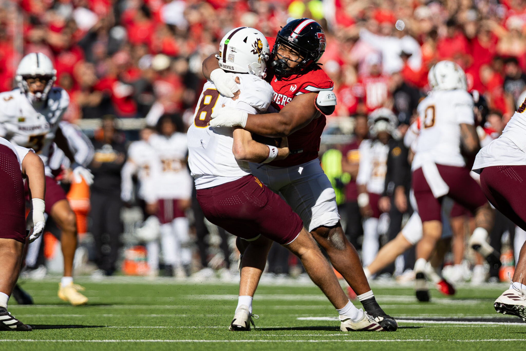 Two football players collide in a tackle on a sunny sideline, one in a red jersey and the other in a white/maroon uniform, with a cheering crowd behind them.