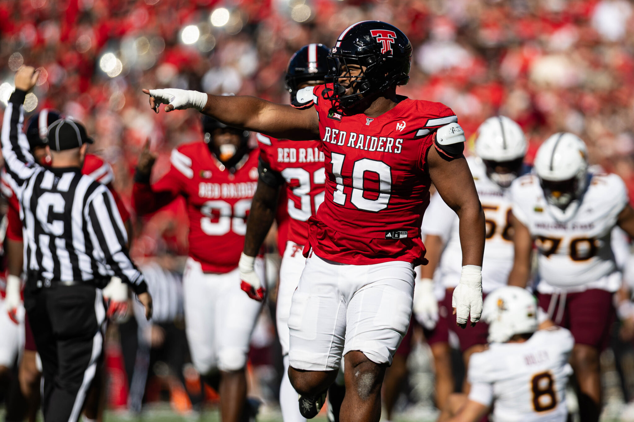 Red Raiders football player in a red jersey and helmet points forward on the field during a game, with a referee and teammates in the background.