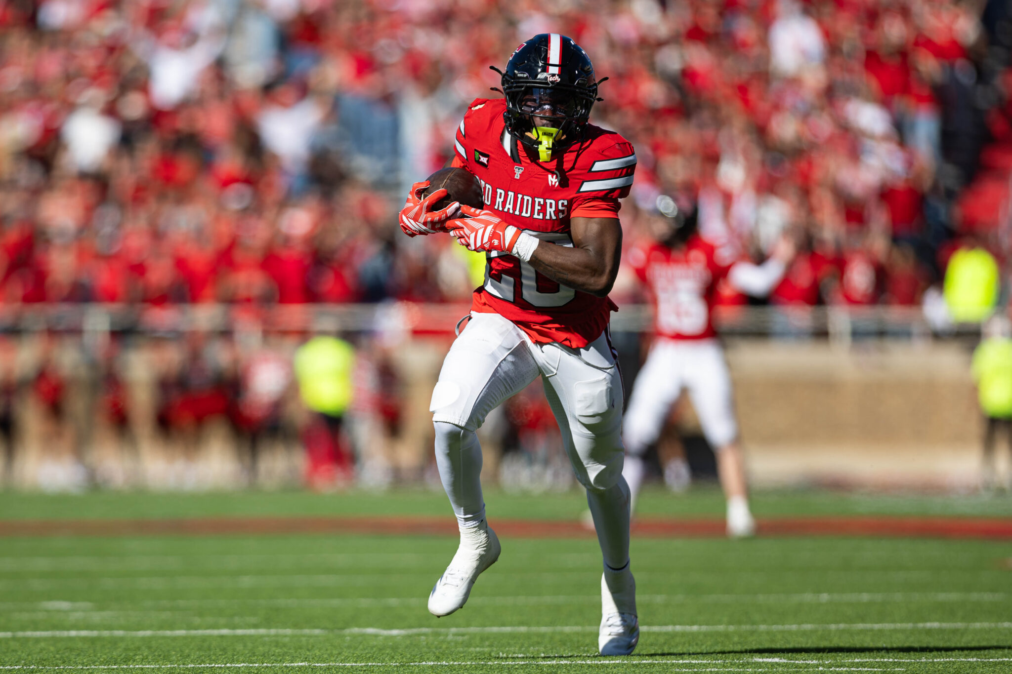 Football player in red Texas Tech/Red Raiders jersey runs with ball during a game, crowd blurred in background.