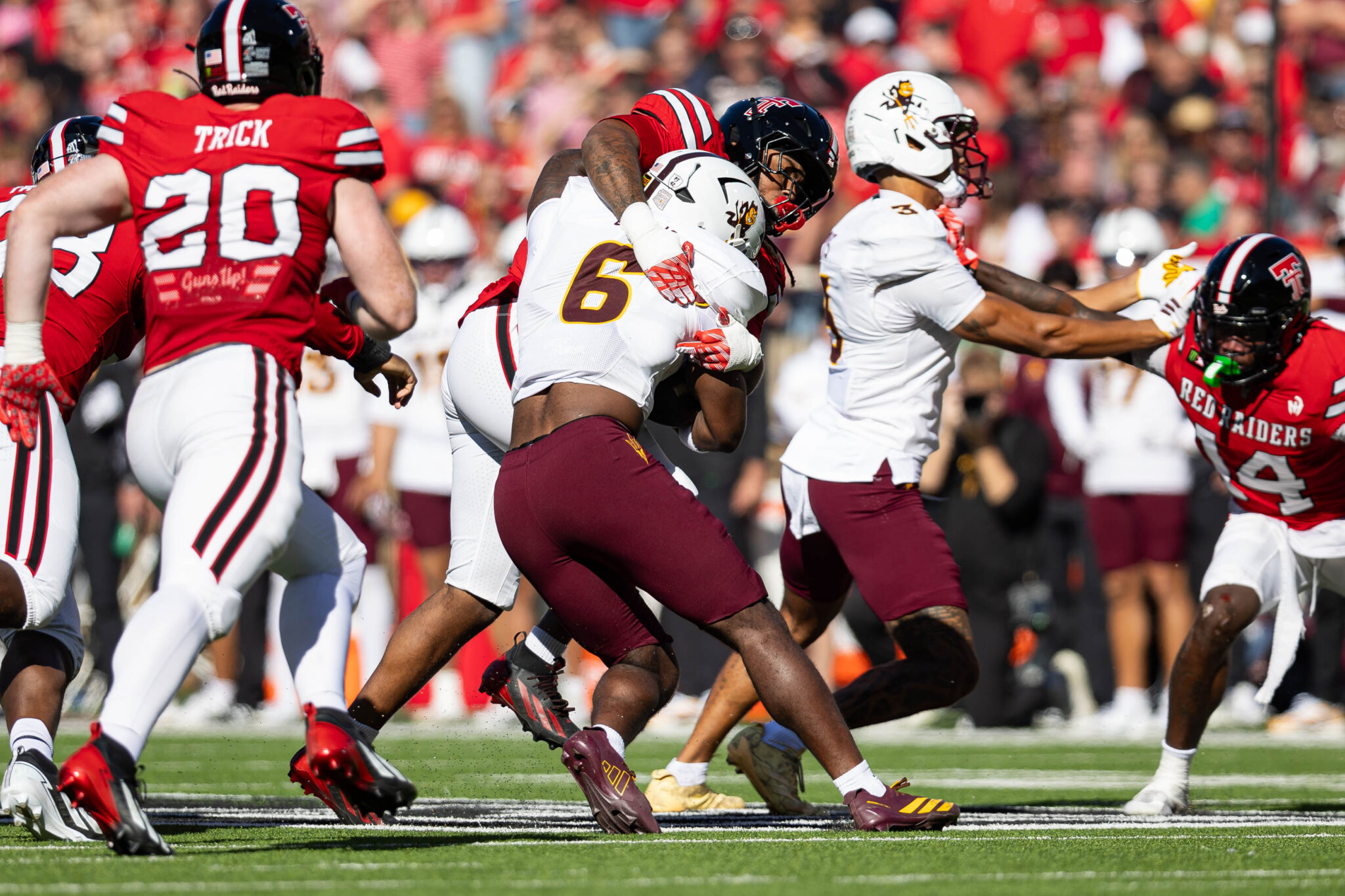 Football players in red and white uniforms clash on the field as the ball is fought for amid a cheering crowd in the stands.