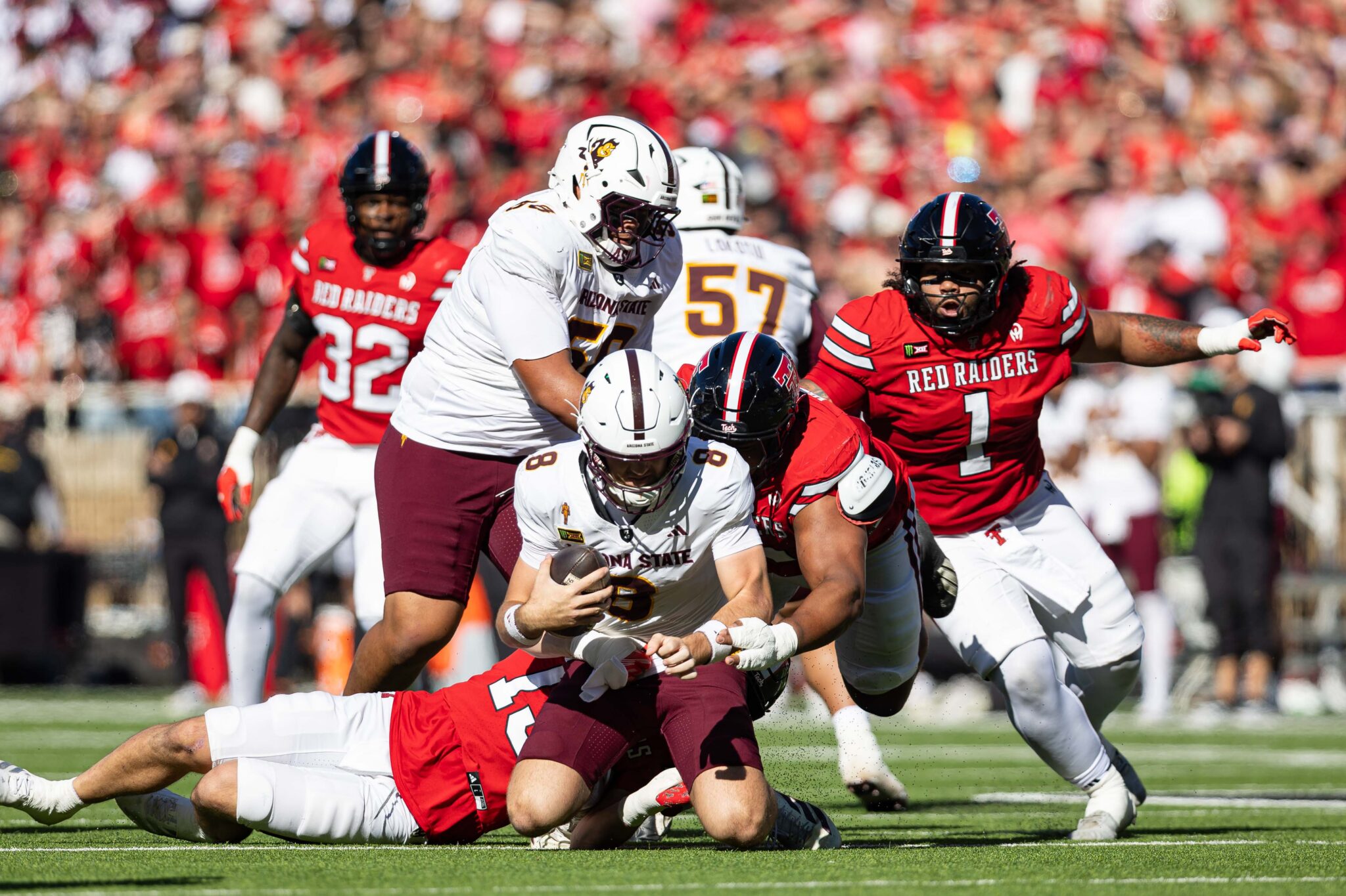 Arizona State ball carrier in white uniform runs with the ball while Red Raiders players in red tackle during a crowded college football game crowd in red stands behind.