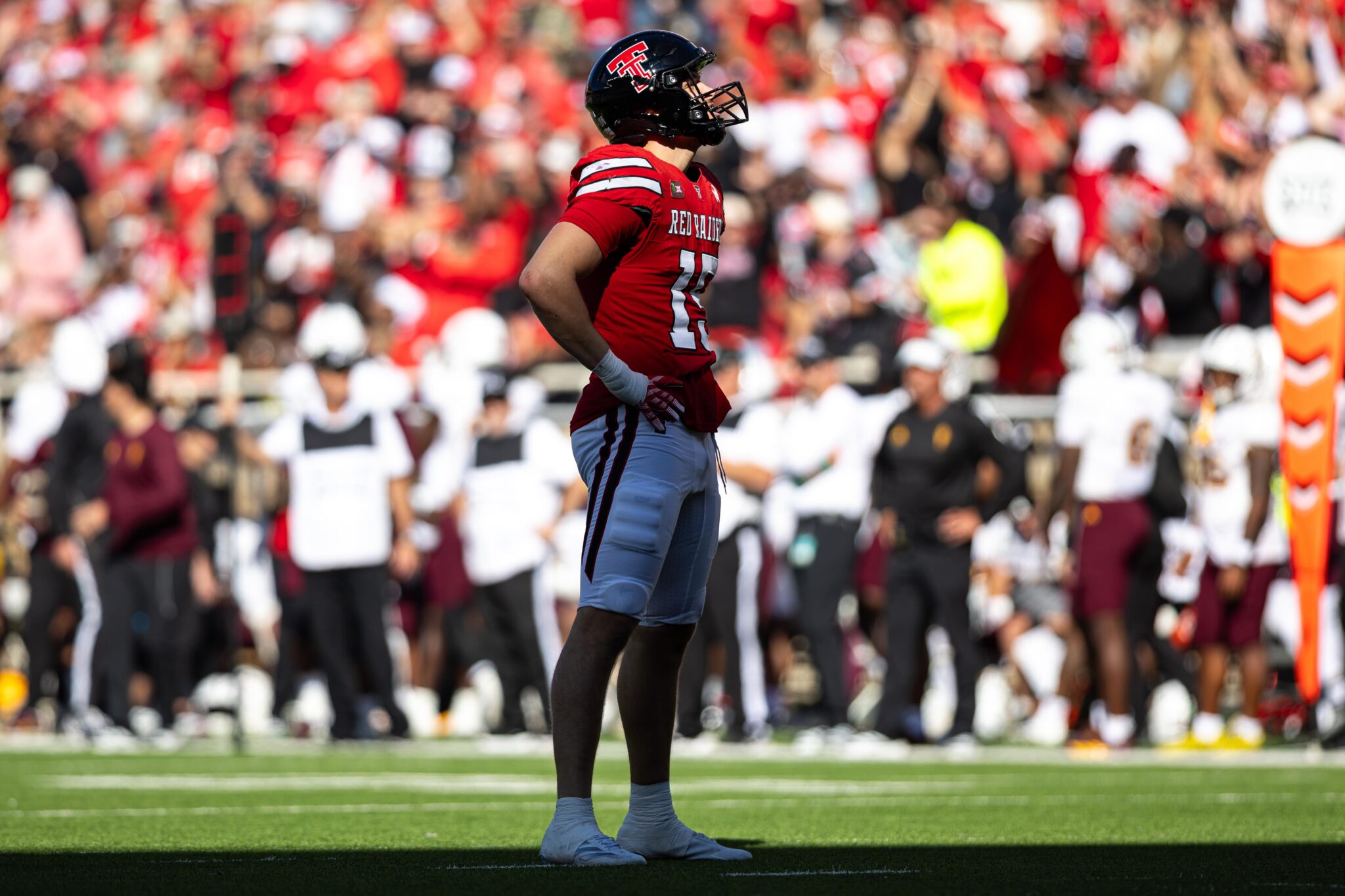 Football player in a red jersey (number 21) and black helmet stands on a green field with a cheering crowd in the background behind him at a stadium.