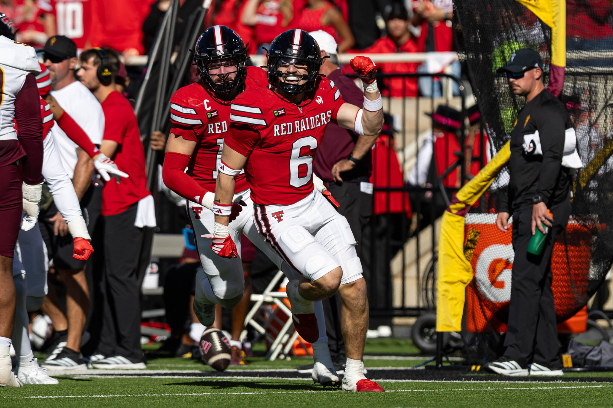 Texas Tech Red Raiders players sprint onto the field in red uniforms, one raising a fist in celebration as fans cheer in red.