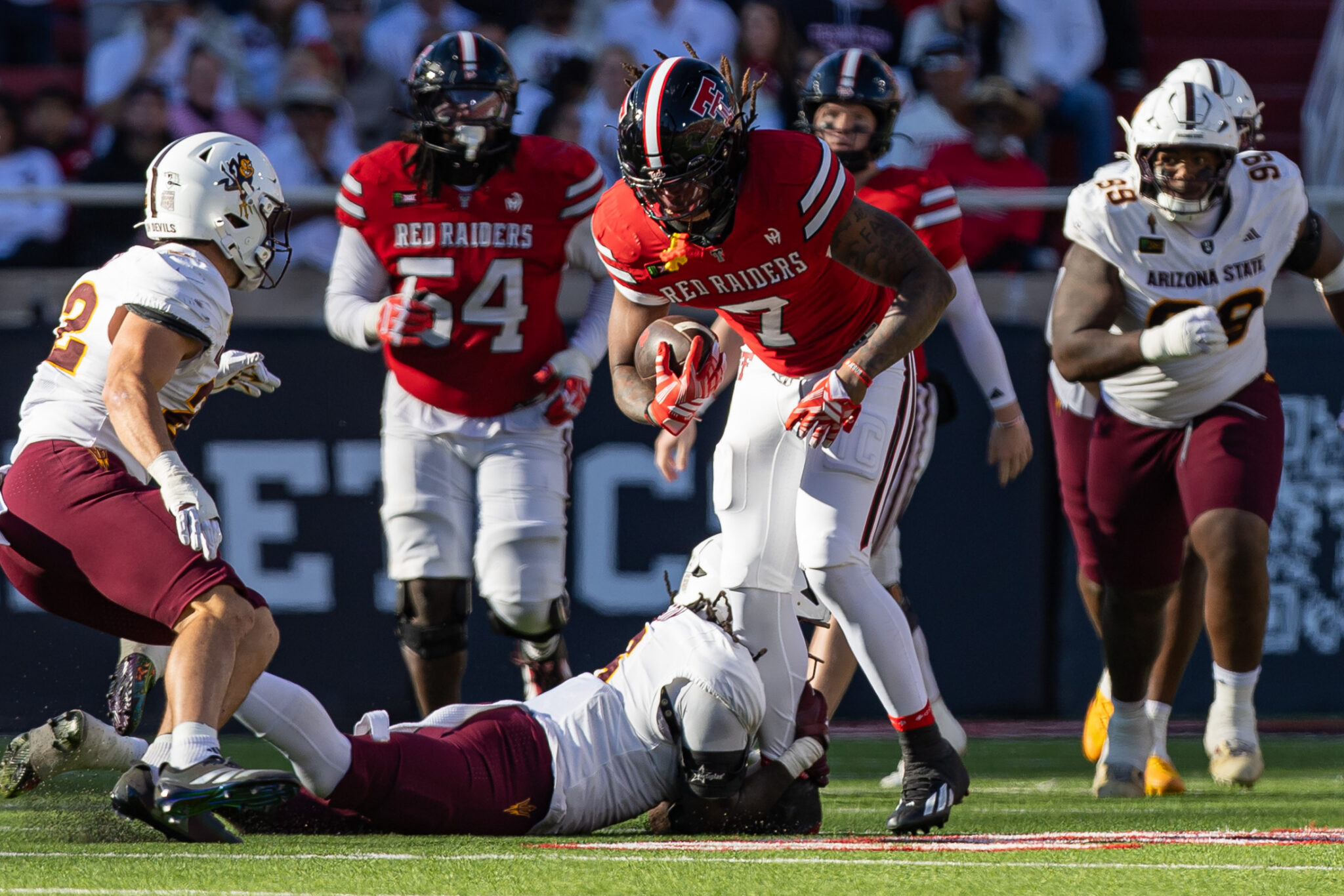 Red Raiders running back with the ball evades defenders during a college football game on a sunlit field, a tackle happening nearby.