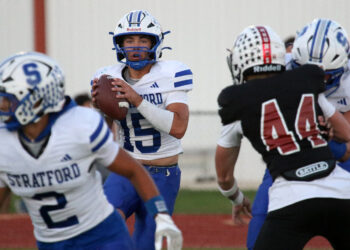 Quarterback in a Stratford white-and-blue uniform grips a football, looking to pass during a football game on a track field.