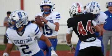 Quarterback in a Stratford white-and-blue uniform grips a football, looking to pass during a football game on a track field.
