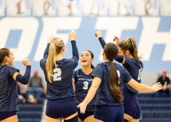 Women’s volleyball team in navy uniforms celebrate with raised arms in a gym.