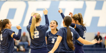 Women’s volleyball team in navy uniforms celebrate with raised arms in a gym.