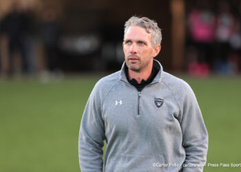 20261015-Dumas-Randall-FB-001 | Press Pass Sports Man wearing a gray Raiders jacket stands on a grassy field, looking off to the right with a neutral expression.