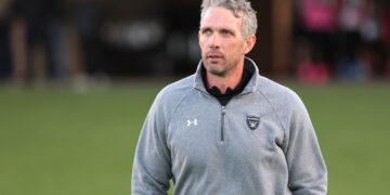 Man wearing a gray Raiders jacket stands on a grassy field, looking off to the right with a neutral expression.