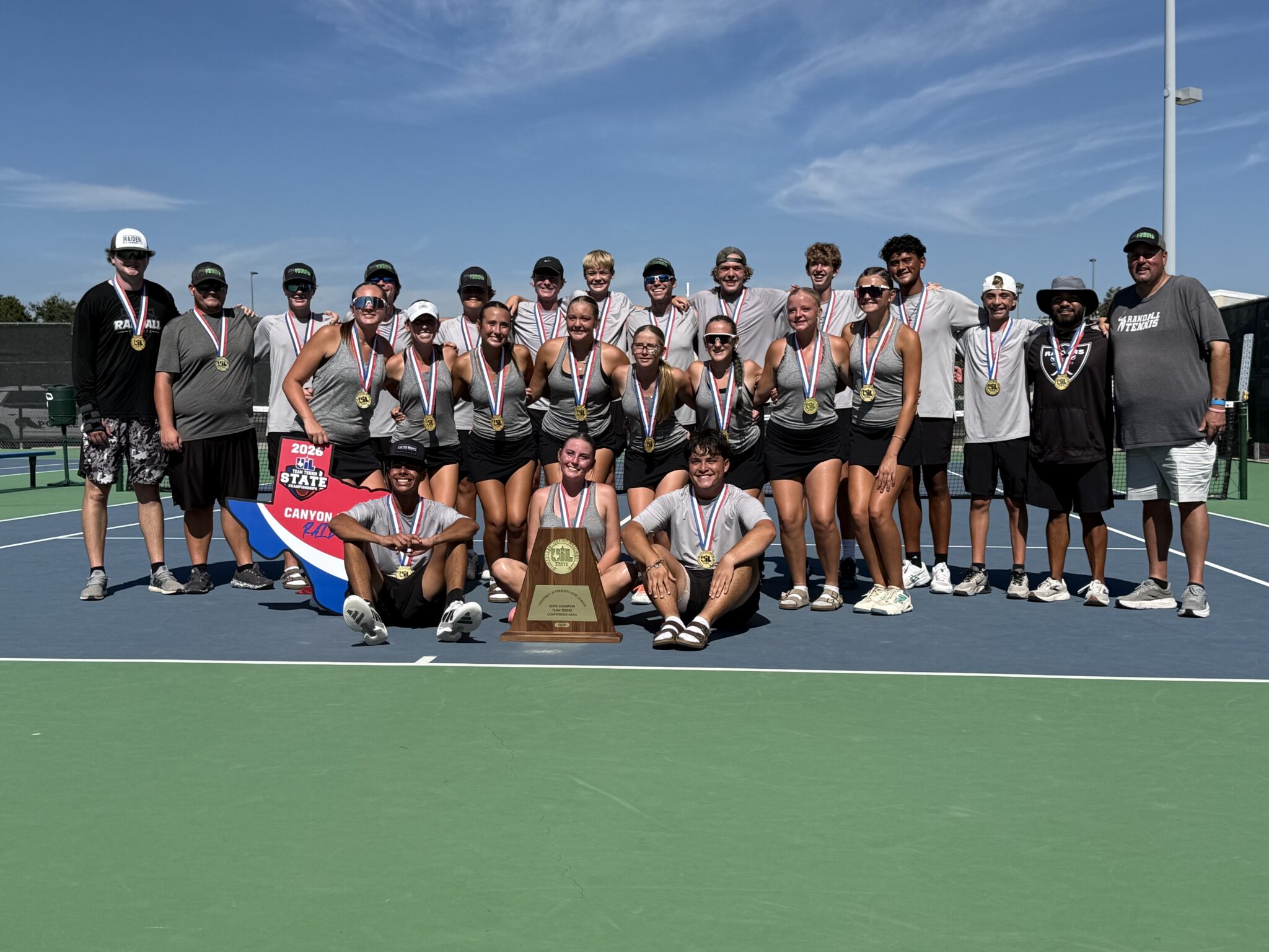 IMG_0978 | Press Pass Sports Group of young tennis players and coaches wearing medals, posing with a trophy on a sunny tennis court.